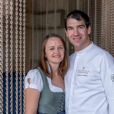Paar vor Kettenvorhang, Mann im Kochoutfit mit Riederalm-Logo, Frau in traditionellem Dirndl